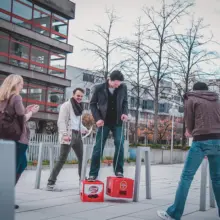 Mann läuft mit Bierkästen als Stelzen. Andere stehen klatschend daneben