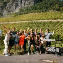 jubelnde Gruppe bei einer Weinwanderung. Neben Ihnen steht ein Bollerwagen. Im Hintergrund die Weinberge