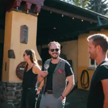 Drei Menschen stehen vor einem Weingut. Ein Mann hat ein Weinglas in der Hand und lächelt in die Kamera